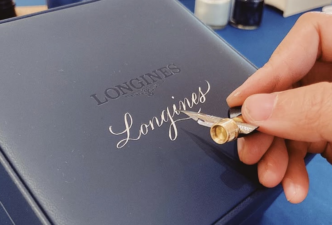 以沾水筆在 Longines 深藍色錶盒上書寫銀色花體字
