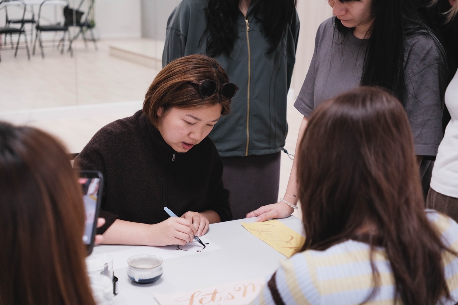 Miko 低頭示範書寫花體字，學員在旁圍觀