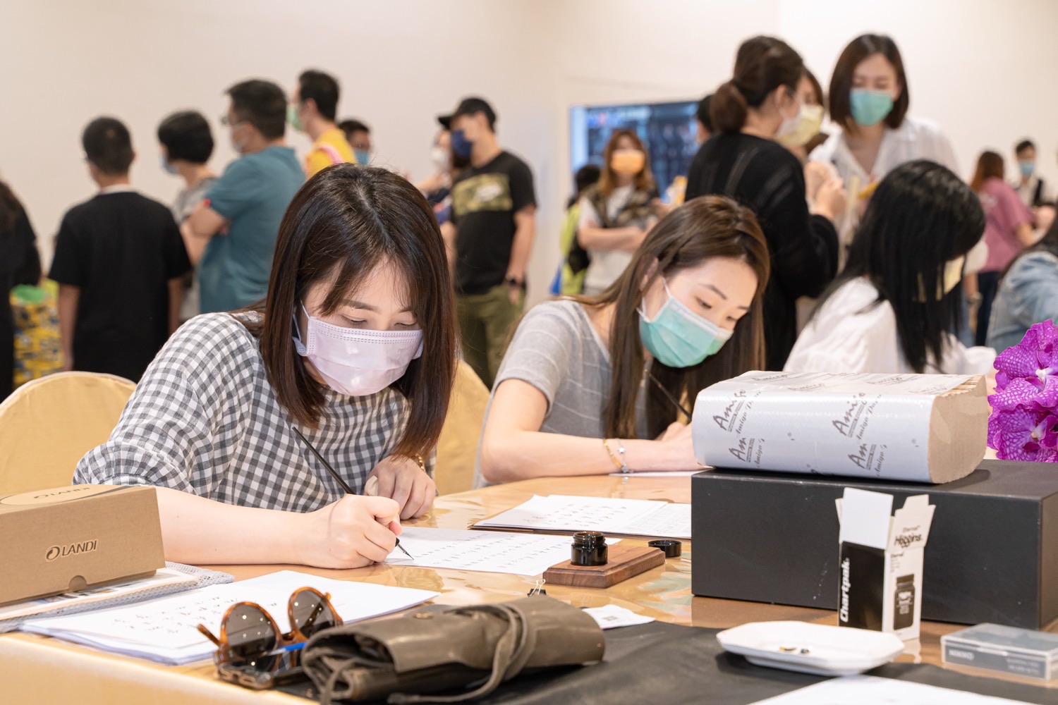 活動現場兩位學員專注書寫，背景人潮往來