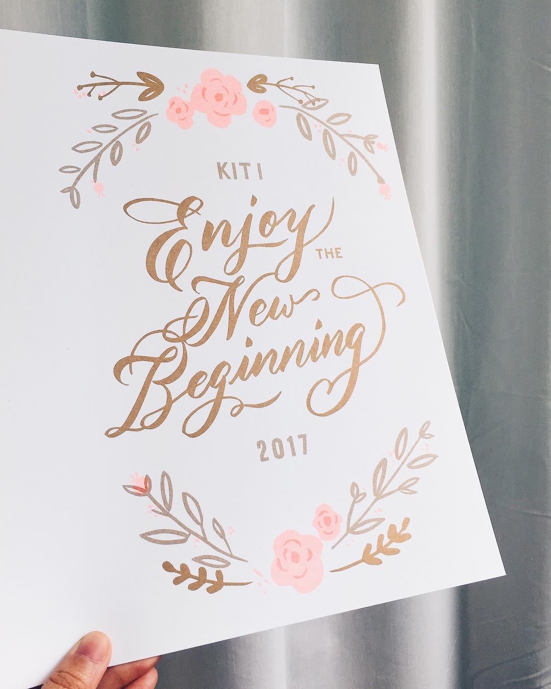以金色花體字書寫 Enjoy the New Beginning 並繪有粉色花環的賀卡